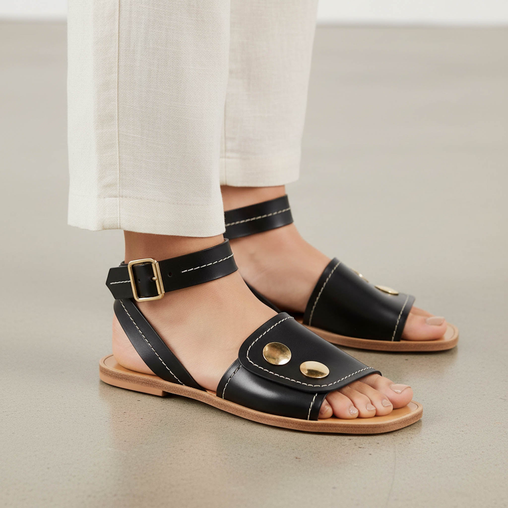 Black sandals with gold accents worn by a person in light-colored pants on a neutral background