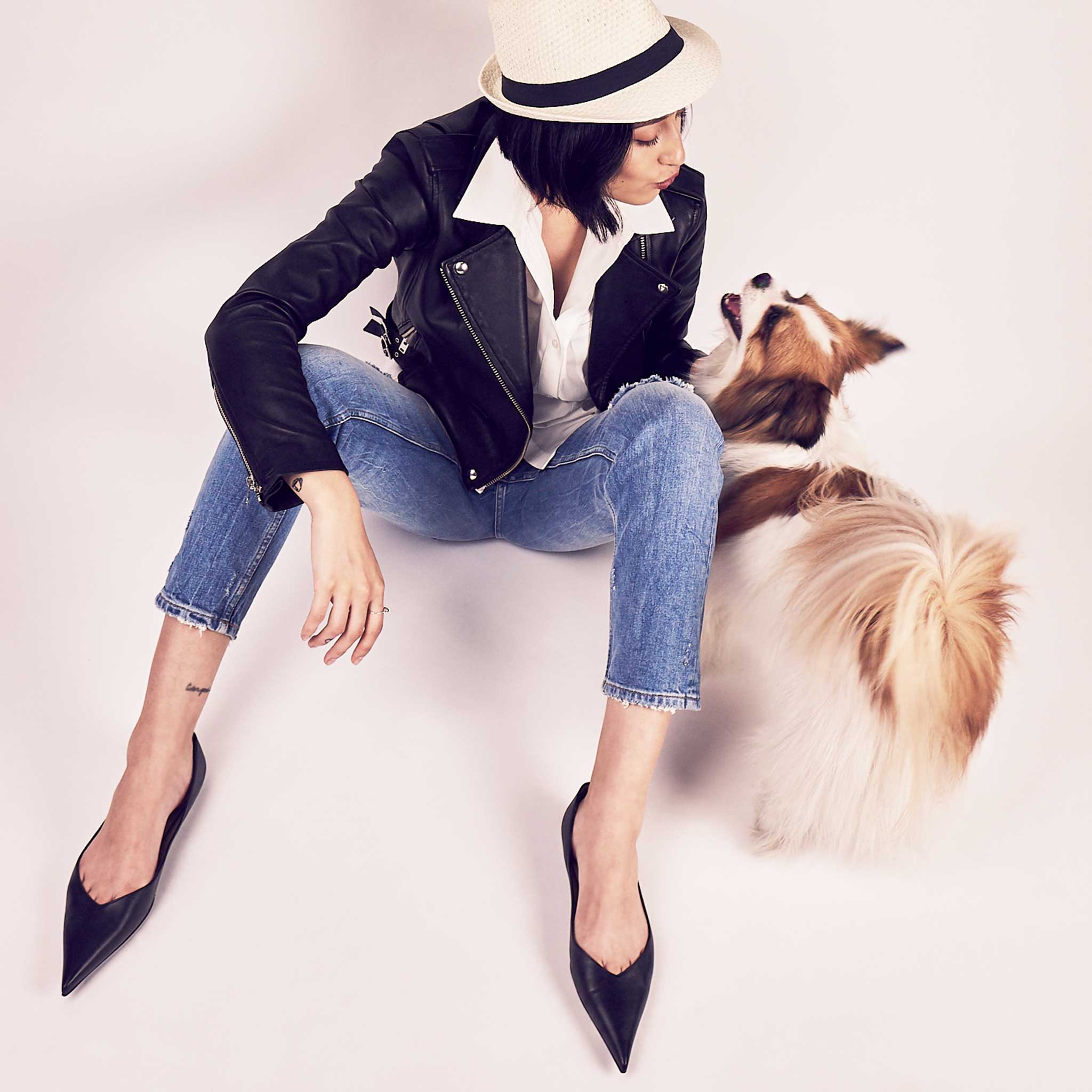 a woman wearing a cream colored fedora, black IRO leather biker jacket, Zara peg leg denim blue jeans and black leather flat shoes with super pointed toe making a kissy face at a small Tibetan spaniel dog who is smiling