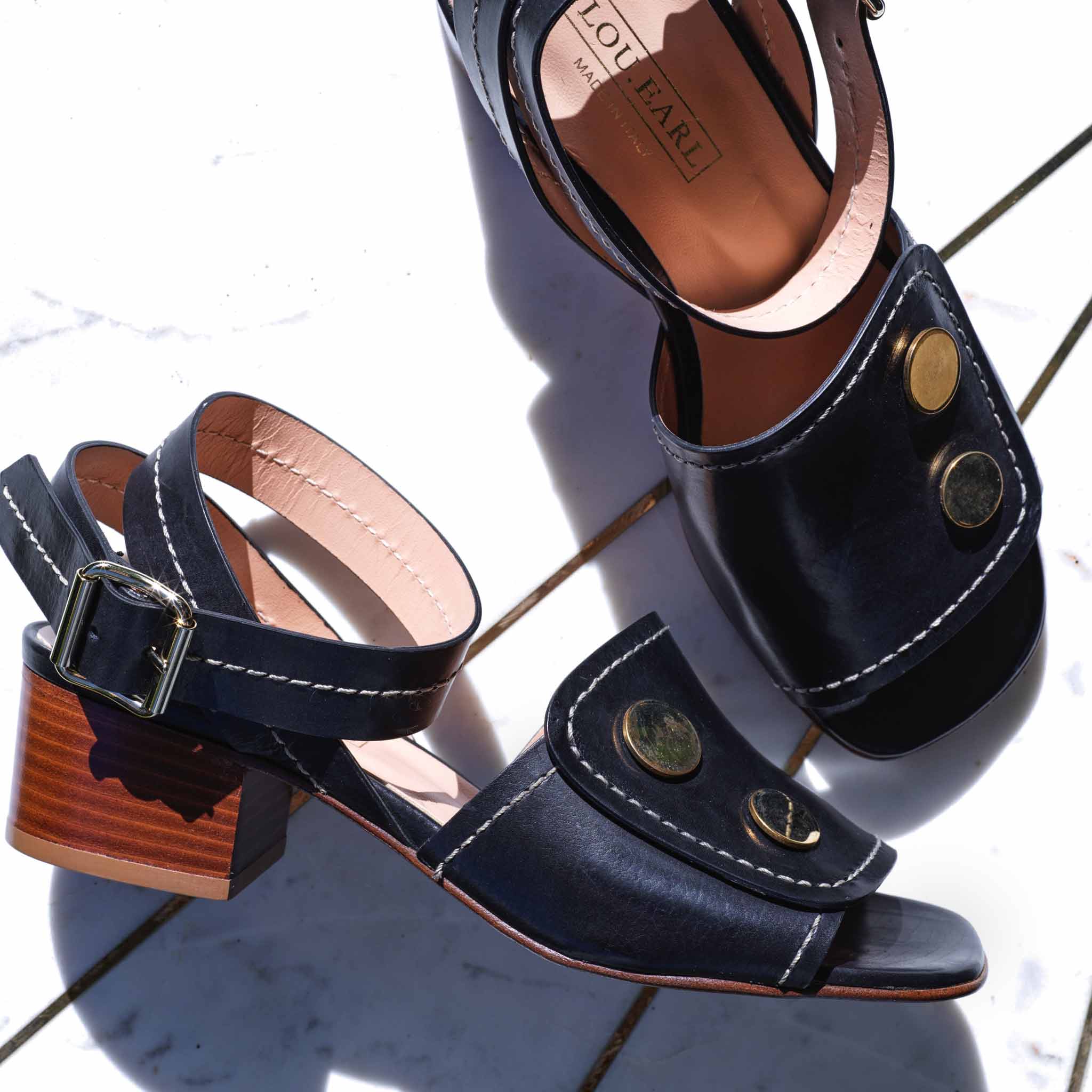 blocky heels with black leather and large round oversized metal studs with white stitching and ankle strap. italian open toe sandals sitting as a pair on marble background.