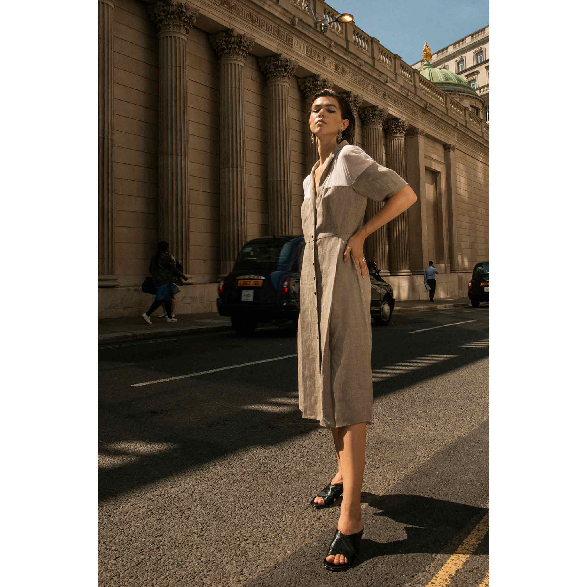 A tall brunette female model standing in London wearing a button up one piece dress and black slip on Lou Earl heeled sandals
