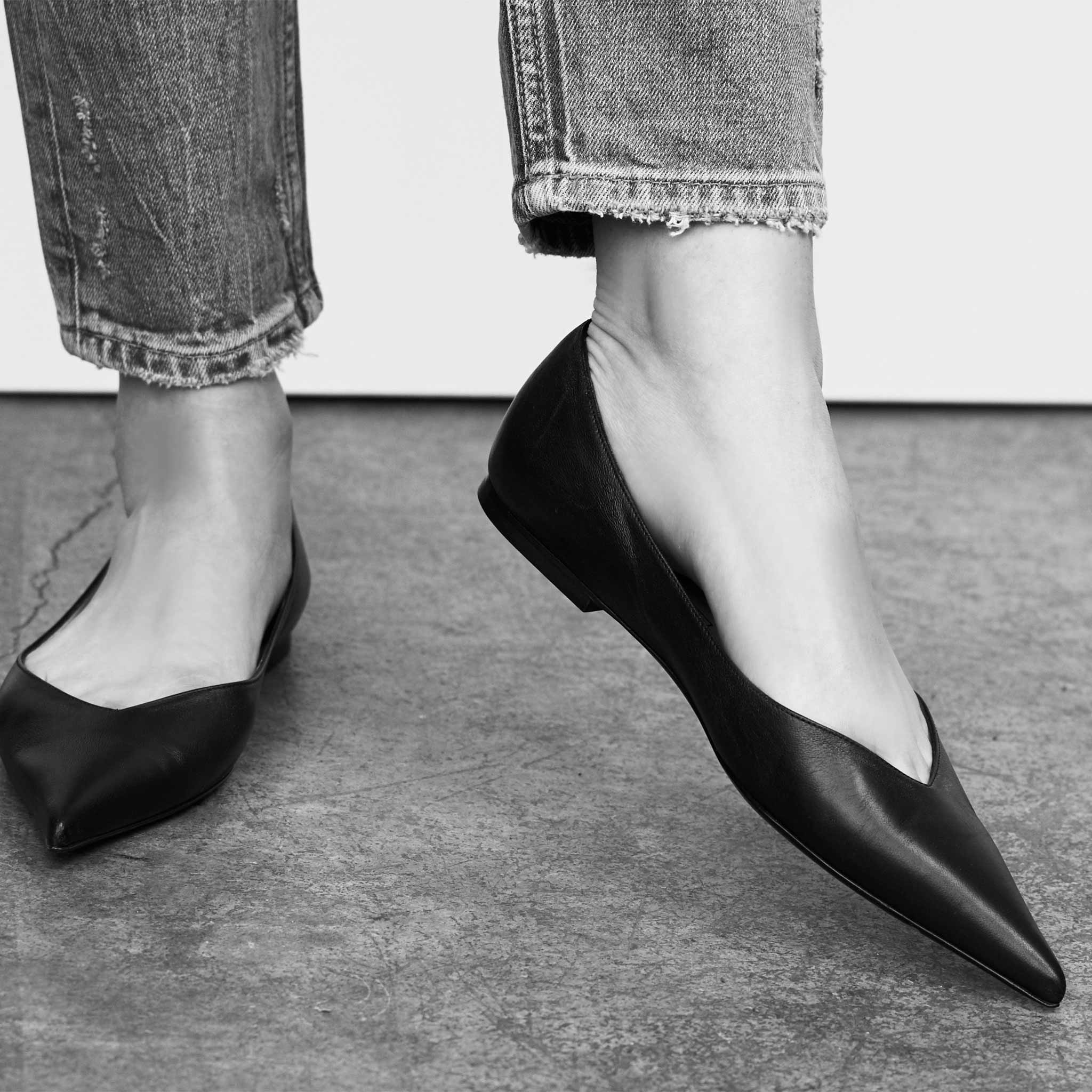 black and white photo of womens feet wearing straight leg distressed denim and black slip on pointed toe skimmer flat shoes